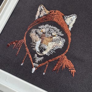 K&ouml;nnte beinhalten: Gestickte Darstellung eines Wolfes, der einen braunen Umhang mit Kapuze tr&auml;gt. Das Fell des Wolfes ist grau und wei&szlig;, mit braunen Akzenten um die Augen und die Nase.