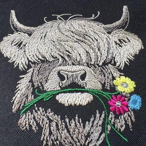May include: Embroidered design of a white Highland cow with long, shaggy hair, holding a bouquet of green leaves and colorful flowers in its mouth. The flowers are yellow, pink, and blue.