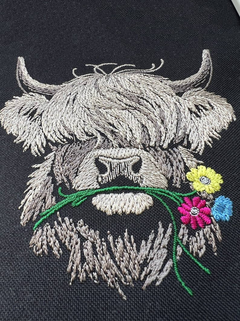 May include: Embroidered design of a white Highland cow with long, shaggy hair, holding a bouquet of green leaves and colorful flowers in its mouth. The flowers are yellow, pink, and blue.