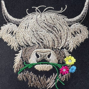 May include: Embroidered design of a Highland cow with long, shaggy hair, brown eyes, and a pink nose. The cow is eating a green stem with pink, yellow, and blue flowers.