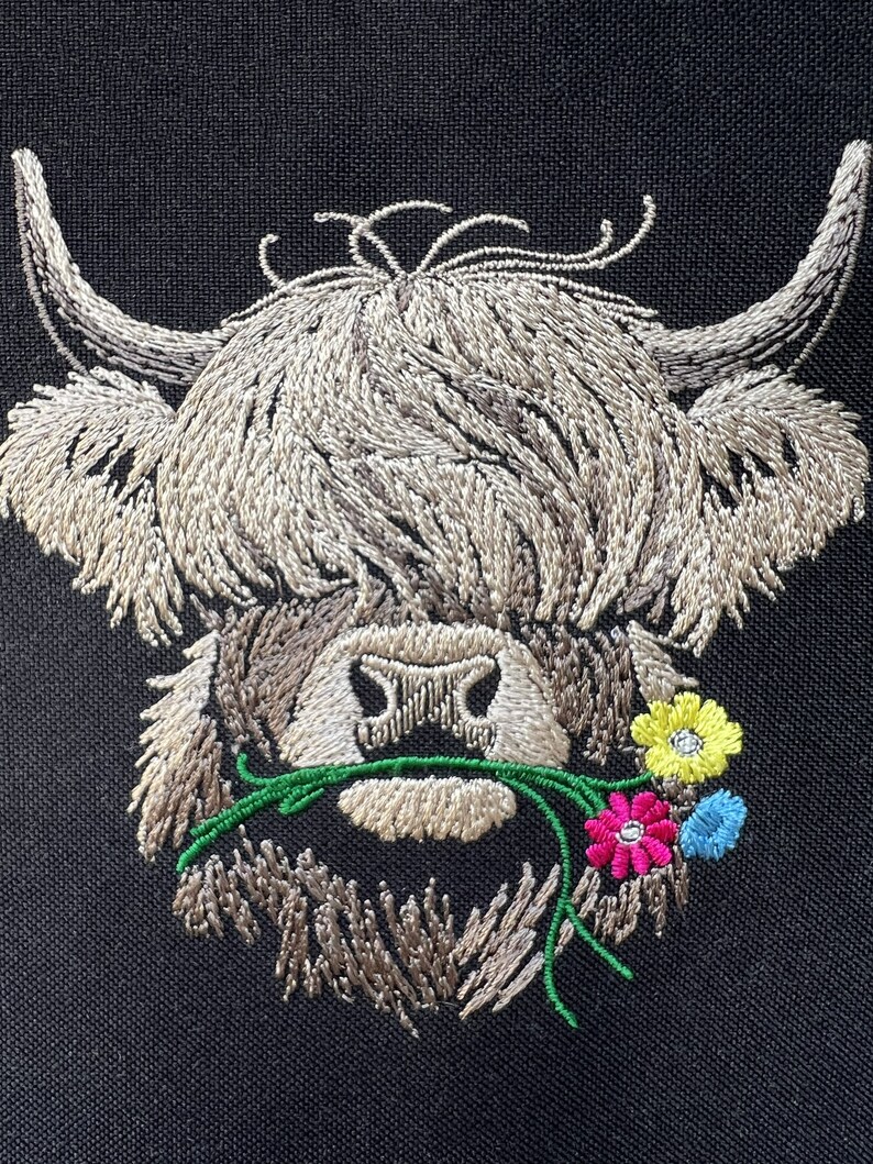 May include: Embroidered design of a Highland cow with long, shaggy hair, brown eyes, and a pink nose. The cow is eating a green stem with pink, yellow, and blue flowers.