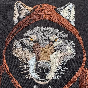 K&ouml;nnte beinhalten: Gestickte Wolfskopf-Darstellung in braunen, grauen und wei&szlig;en Farben auf einem schwarzen Stoffhintergrund. Das Wolfsgesicht ist von einem braunen Kreisrahmen umgeben.