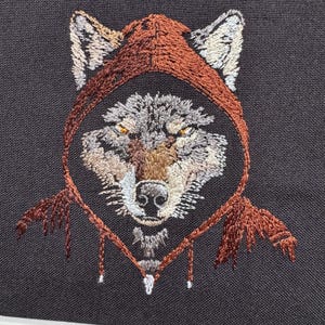 K&ouml;nnte beinhalten: Gestickte Darstellung eines Wolfskopfes, der einen braunen Kapuzenmantel tr&auml;gt. Das Fell des Wolfes ist grau und braun, mit einer wei&szlig;en Schnauze und gelben Augen.
