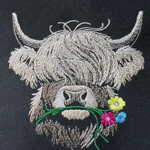 May include: Embroidered design of a Highland cow with brown fur, a white face, and a green stem with pink, yellow, and blue flowers in its mouth.