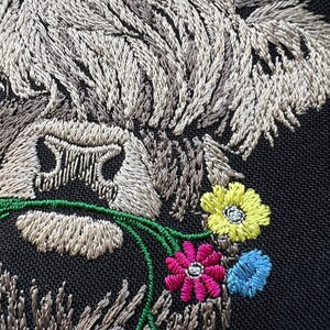 May include: Embroidered patch of a Highland cow with a white and gray fur pattern, looking to the right, with its head tilted down. The cow is eating a bouquet of green, pink, yellow, and blue flowers.