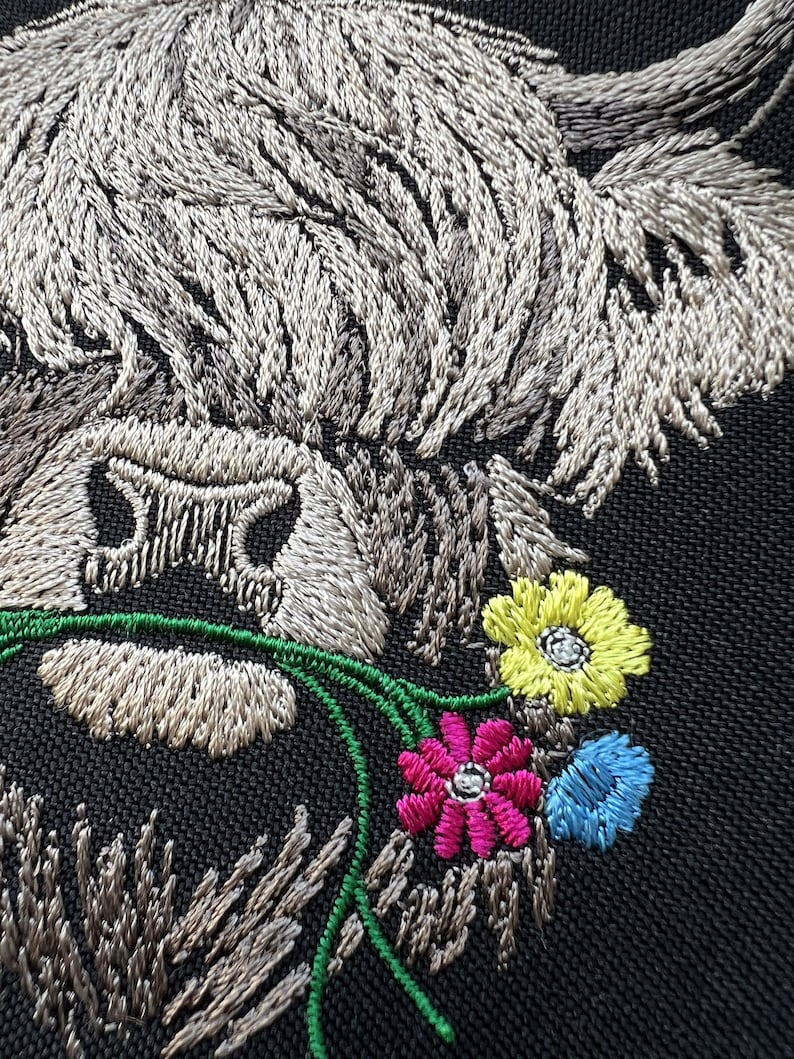 May include: Embroidered patch of a Highland cow with a white and gray fur pattern, looking to the right, with its head tilted down. The cow is eating a bouquet of green, pink, yellow, and blue flowers.