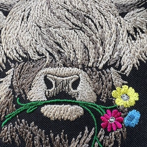 May include: Embroidered design of a Highland cow with a fluffy brown and white coat, holding a bouquet of colorful flowers in its mouth. The flowers are pink, yellow, blue, and green.