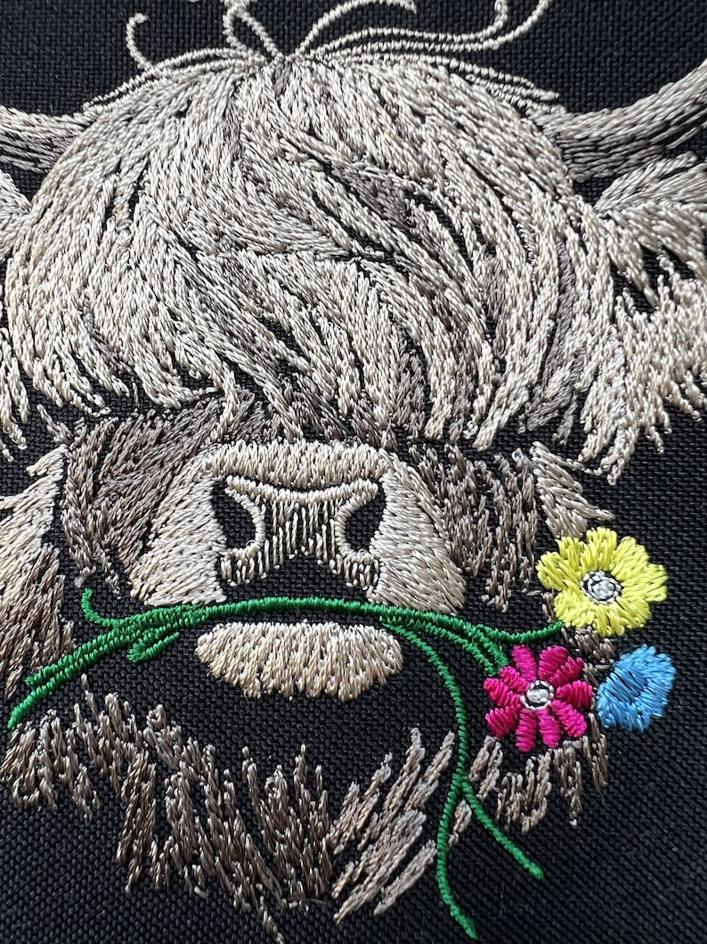 May include: Embroidered design of a Highland cow with a fluffy brown and white coat, holding a bouquet of colorful flowers in its mouth. The flowers are pink, yellow, blue, and green.
