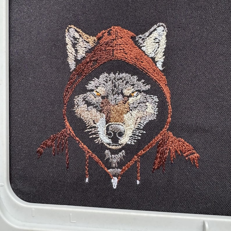 K&ouml;nnte beinhalten: Gestickte Darstellung eines Wolfes, der einen roten Umhang mit Kapuze tr&auml;gt. Das Fell des Wolfes ist grau und braun, und seine Augen sind gelb. Das Design ist auf einem schwarzen Stoffhintergrund.