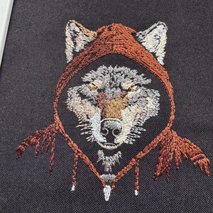 K&ouml;nnte beinhalten: Gestickte Darstellung eines Wolfes, der einen braunen Kapuzenmantel tr&auml;gt. Das Fell des Wolfes ist grau und braun, mit einer wei&szlig;en Schnauze und gelben Augen.
