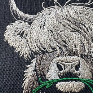 May include: Embroidered image of a Highland cow's head with a long, white horn and a fluffy white face. The cow is eating green grass.