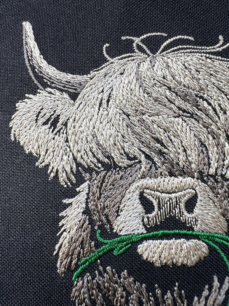 May include: Embroidered image of a Highland cow's head with a long, white horn and a fluffy white face. The cow is eating green grass.
