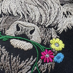 May include: Embroidered design of a Highland cow with a white and gray fur, holding a bouquet of green, pink, yellow, and blue flowers.