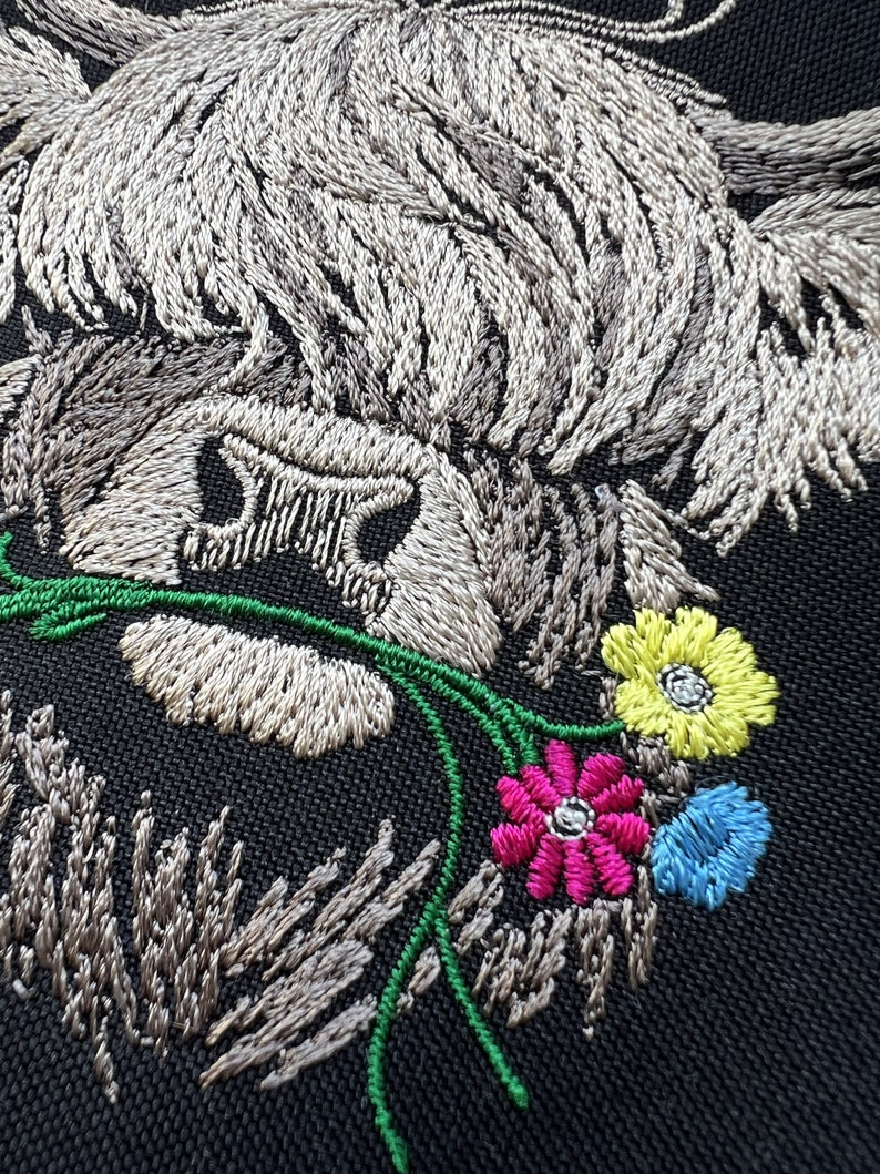 May include: Embroidered design of a Highland cow with a white and gray fur, holding a bouquet of green, pink, yellow, and blue flowers.