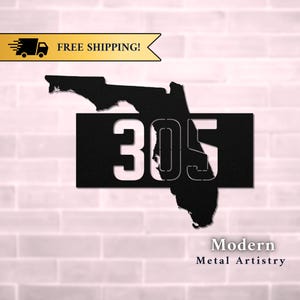 May include: Black metal house number sign with the number 305 cut out. The number is centered on a black rectangle with the outline of the state of Florida cut out behind it. The sign is on a white brick wall. Modern Metal Artistry.