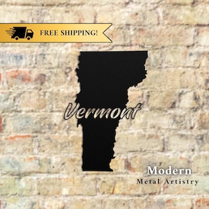 May include: A black metal cutout of the state of Vermont, with the state's name in a cursive font. The words "Modern Metal Artistry" are below the state outline. The background is a brick wall.