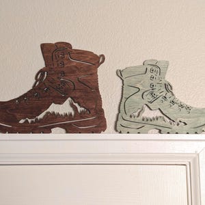 May include: Two wooden hiking boot wall decor pieces. The boot on the left is brown and the boot on the right is light green. Both boots have a mountain scene cut out of the wood.