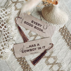 May include: Two rectangular bookmarks with brown tassels. The top bookmark reads "EVERY GOOD BOOK..." with a horseshoe graphic. The bottom bookmark reads "...HAS A COWBOY IN IT" with a cowboy hat graphic. The bookmarks are on a textured surface.