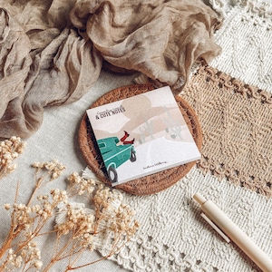 May include: A notepad featuring the text "Country Roads & Cute Notes" and an illustration of a teal truck with a person's legs visible. The notepad sits on a woven coaster, with neutral-toned fabric and dried flowers in the background.