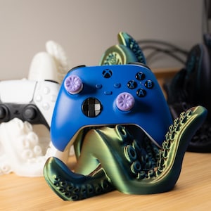 May include: A blue Xbox controller rests on a metallic green octopus-shaped stand. A white PlayStation controller and a black octopus stand are also visible. The stands are designed to hold gaming controllers.