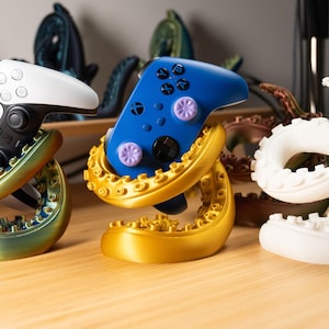 Op de afbeelding: Drie gamecontroller-standaards in de vorm van octopus-tentakels. Eén standaard houdt een wit en zwarte controller vast, een andere een blauwe controller en de derde is wit. De standaards zijn goud, groen en wit.