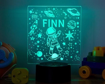 May include: A glowing night light with an astronaut and space-themed illustrations, including rockets, planets, and stars. The name "FINN" is displayed at the top. Colorful toy blocks and a toy car are on either side of the lamp.