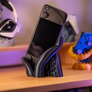 Pode incluir: Um dispositivo de jogos portátil preto é segurado por um suporte preto em forma de mão impresso em 3D. O suporte está sobre uma superfície de madeira. Outros acessórios de jogos são visíveis no fundo.