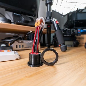 May include: An electrical component with red and black wires, a black mounting base, and a separate black ring. The component is on a wooden surface. The wires are connected to a clear plastic connector.