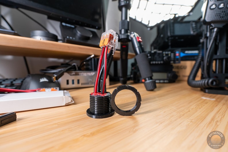May include: An electrical component with red and black wires, a black mounting base, and a separate black ring. The component is on a wooden surface. The wires are connected to a clear plastic connector.