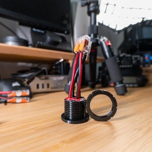 May include: A black electrical component with red and black wires extending from it. A black circular ring sits next to the component. The items are on a wooden surface. The image is well-lit and in focus.