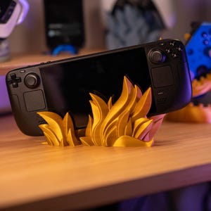 May include: A black handheld gaming console sits on a gold-coloured flame-shaped stand. The stand is 3D-printed and placed on a wooden surface. Other gaming accessories are visible in the background.