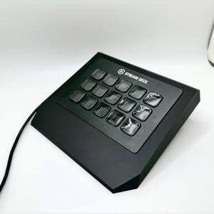 May include: Black Stream Deck with 15 illuminated buttons. The device has the text "STREAM DECK" printed on the top. A black cable extends from the device. The device is designed for live streaming and content creation.