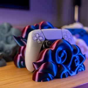 Puede incluir: Un mando blanco y negro de PlayStation 5 descansa en un soporte impreso en 3D. El soporte tiene un diseño en espiral con tonos azules y rojos. Otros soportes de mando son visibles en el fondo.