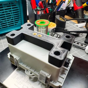 May include: A grey and white electronic component with a black bracket, featuring two gold-coloured connectors. The component is on a black surface, with a teal fan and various tools in the background. The image is taken in a workshop setting.