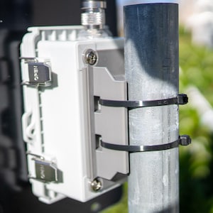 以下が含まれることがあります： 白とグレーの電子機器が、黒い結束バンドで金属製のポールに取り付けられています。 このデバイスには銀色のコネクタがあり、黒い金属板に取り付けられています。 「OPEN」の文字がラッチに見えます。