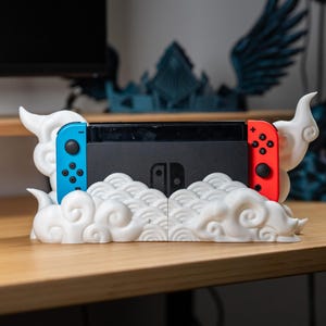 May include: A white 3D-printed cloud-shaped stand holding a black Nintendo Switch console with blue and red Joy-Con controllers. The stand is on a wooden surface, with decorative objects in the background.