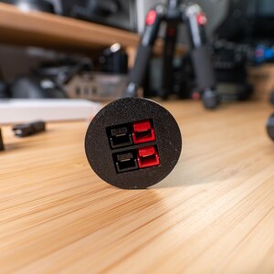 May include: Black circular electrical connector with red and black terminals. The connector is on a wooden surface, with other electronic components in the background. The connector has two pairs of terminals.