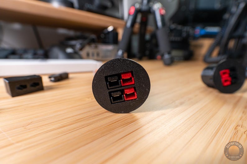May include: Black circular electrical connector with red and black terminals. The connector is on a wooden surface, with other electronic components in the background. The connector has two pairs of terminals.