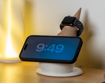 May include: A black smartphone displaying the time 9:49 rests on a white wireless charging stand. A black smartwatch sits atop a brown conical stand. The setup is on a wooden surface, with a glowing lamp in the background.