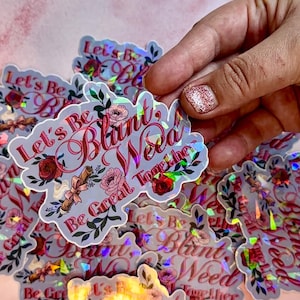 May include: Holographic sticker with the text "Let's Be Blunt, Weed Together" in a cursive font, decorated with floral and leaf illustrations. The sticker is held by a hand with glitter nail polish.