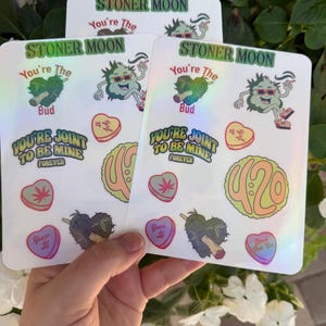 May include: Holographic stickers with cannabis-themed designs. The stickers feature a cartoon bud character, hearts, and the phrases "STONER MOON", "You're The Bud", "YOU'RE JOINT TO BE MINE FOREVER", and "420".