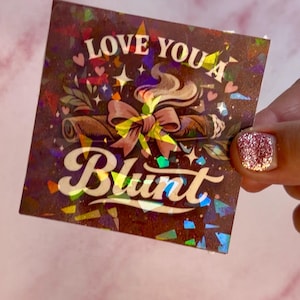 May include: A holographic sticker with the words "LOVE YOU A Blunt". The sticker features an illustration of a rolled item with smoke, a bow, hearts, and stars. The background is a dark brown colour.