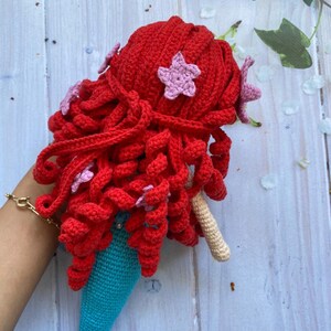 May include: A crocheted mermaid doll with red hair and a turquoise tail. The doll is decorated with pink star-shaped embellishments.