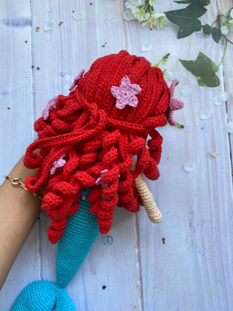 May include: A crocheted mermaid doll with red hair and a turquoise tail. The doll is decorated with pink star-shaped embellishments.