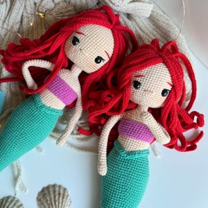May include: Two crocheted mermaid dolls with red hair and green tails. The dolls are wearing purple tops and have smiling faces.