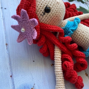 May include: A crocheted doll with red hair, a purple flower, and a blue top. The doll is lying on a blue wooden surface.
