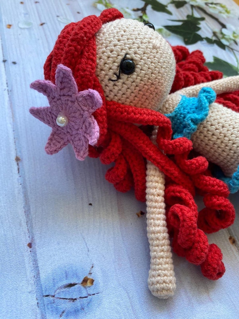 May include: A crocheted doll with red hair, a purple flower, and a blue top. The doll is lying on a blue wooden surface.