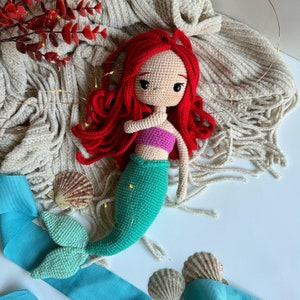 May include: A crocheted mermaid doll with red hair, a pink top, and a green tail. The doll is lying on a white surface with a beige blanket and seashells.
