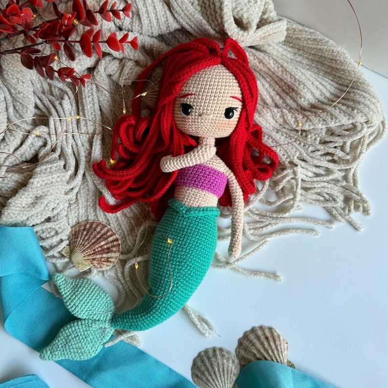 May include: A crocheted mermaid doll with red hair, a pink top, and a green tail. The doll is lying on a white surface with a beige blanket and seashells.