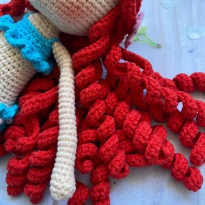 May include: A crocheted doll with long, curly red hair and a blue top. The doll is made with white yarn and has a sweet, innocent expression.